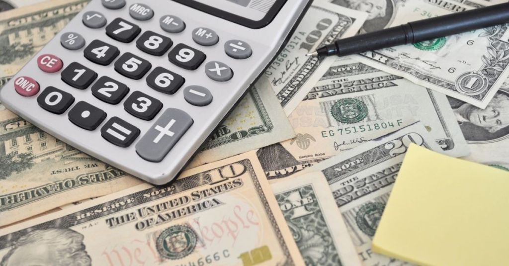 A calculator, sticky notes, and a pen sitting on top of a bed of twenty-dollar bills