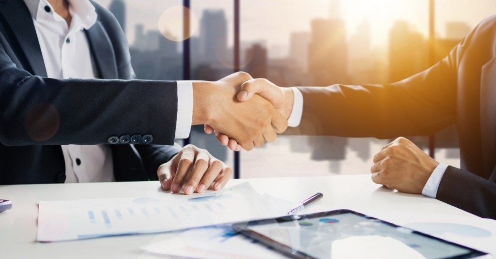 Two men shaking hands in a conference room with a city skyline background.