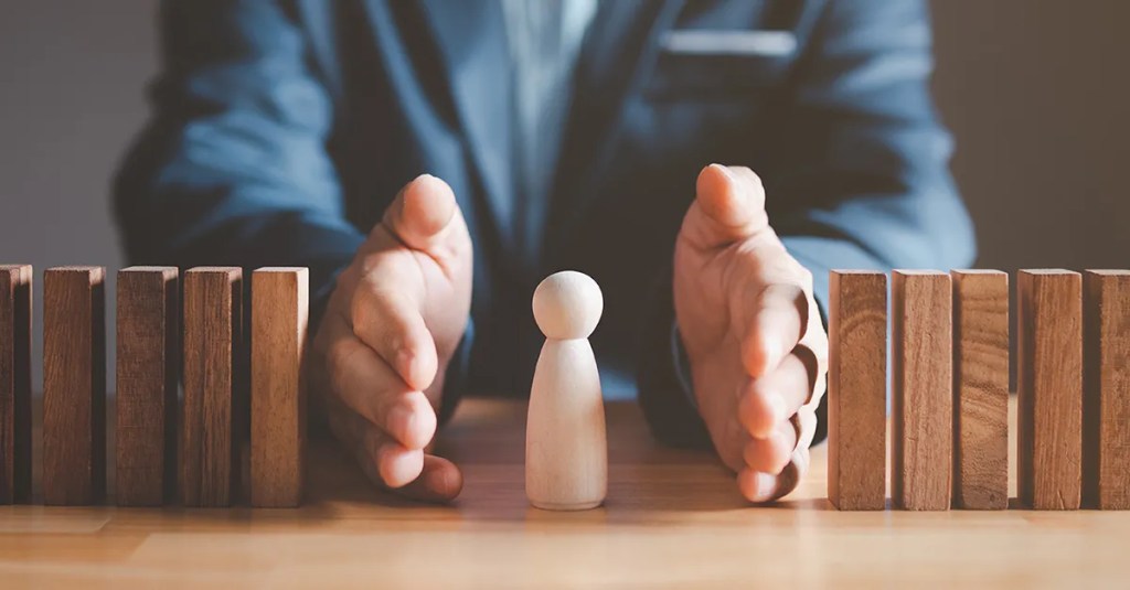 A man protecting a wooden human figurine with his hands from two rows of dominoes.