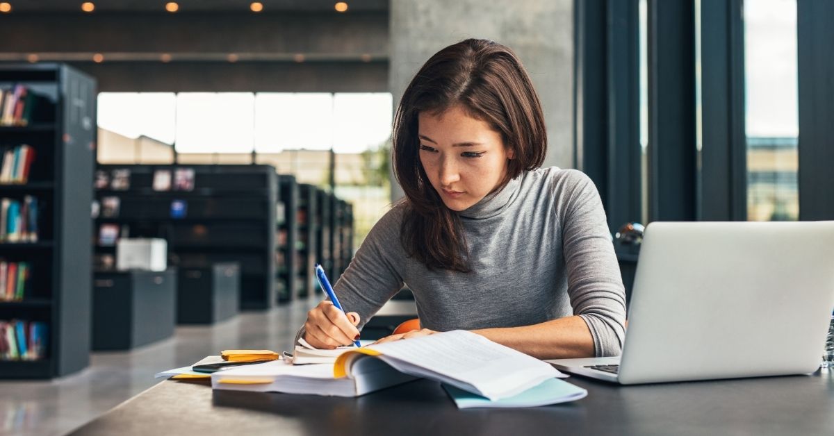 A woman in a library writing in a notebook and looking at her notes.