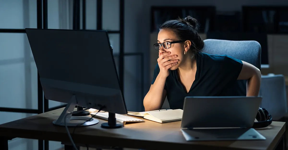 A woman looking shocked with her hand over her mouth, looking at a computer screen in a dark room.