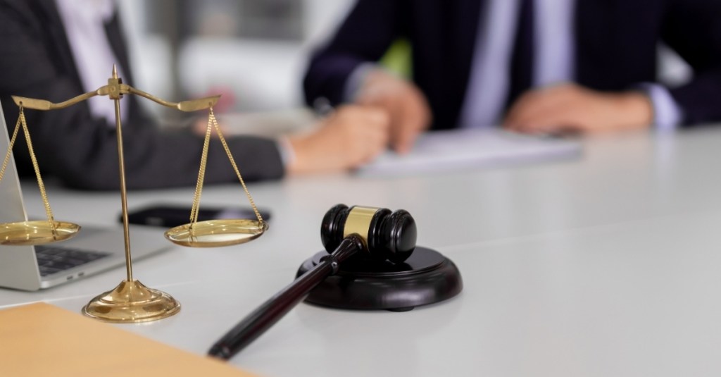 A checks and balances scale and gavel with a blurred background of two men in suits looking over a document.